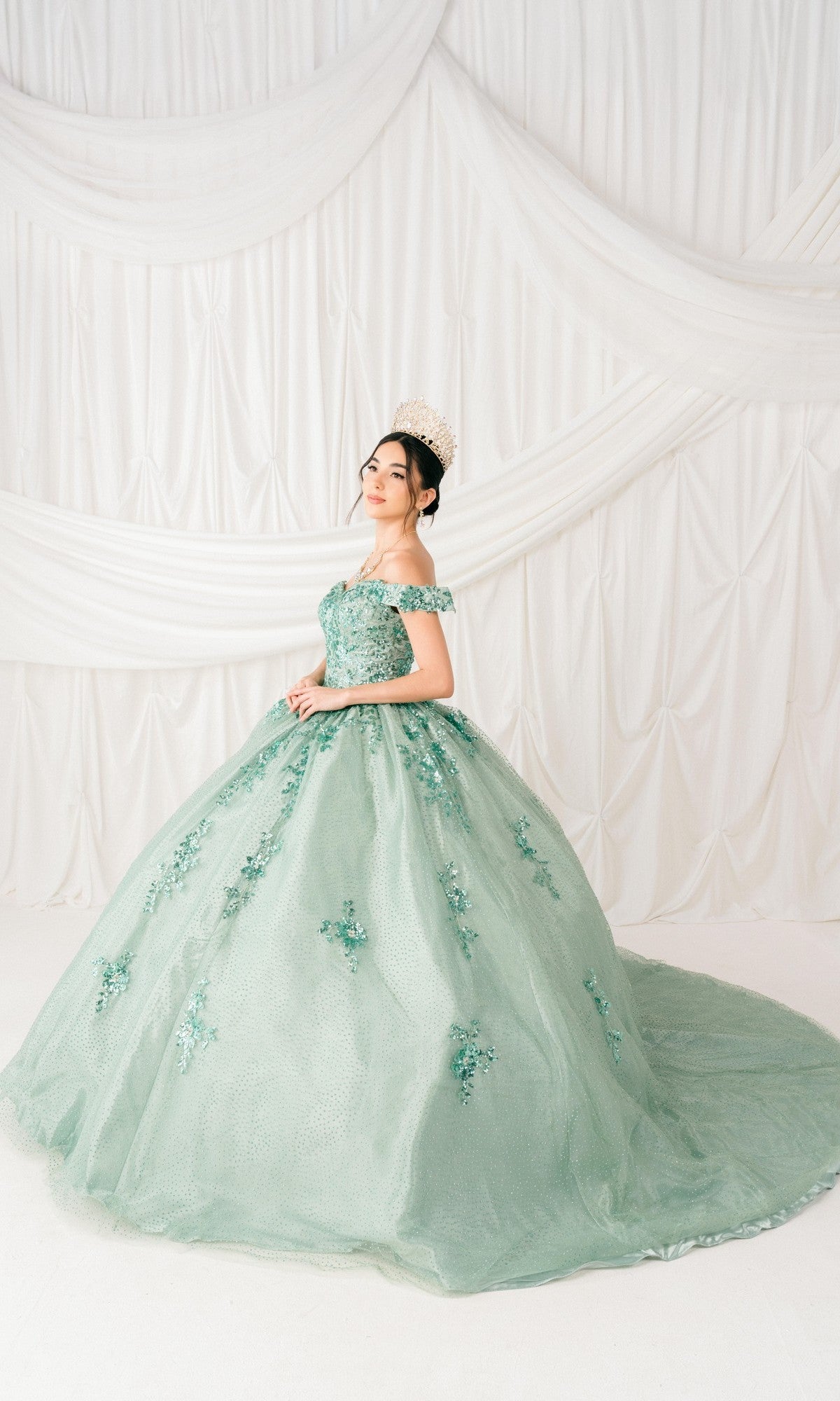 A young woman in the Quinceanera Ball Gown 8060J, featuring embossed tulle and floral details, with a crown, stands before white draped fabric.